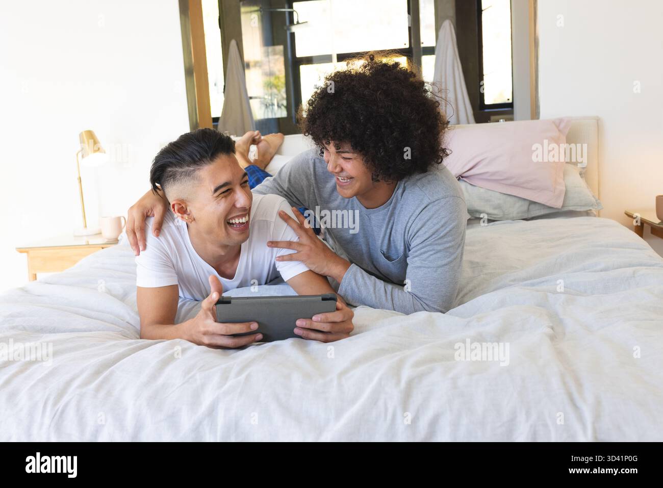 Rire divers amis masculins allongés sur le lit dans une chambre moderne, tenant une tablette près de la lampe et des oreillers Banque D'Images