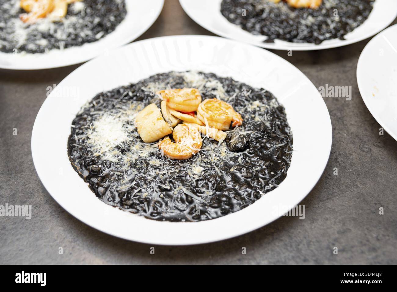 Nourriture, gros plan d'un élégant risotto à l'encre de calmar servi avec des crevettes, des pétoncles et du parmesan sur une assiette blanche dans un cadre de restaurant Banque D'Images