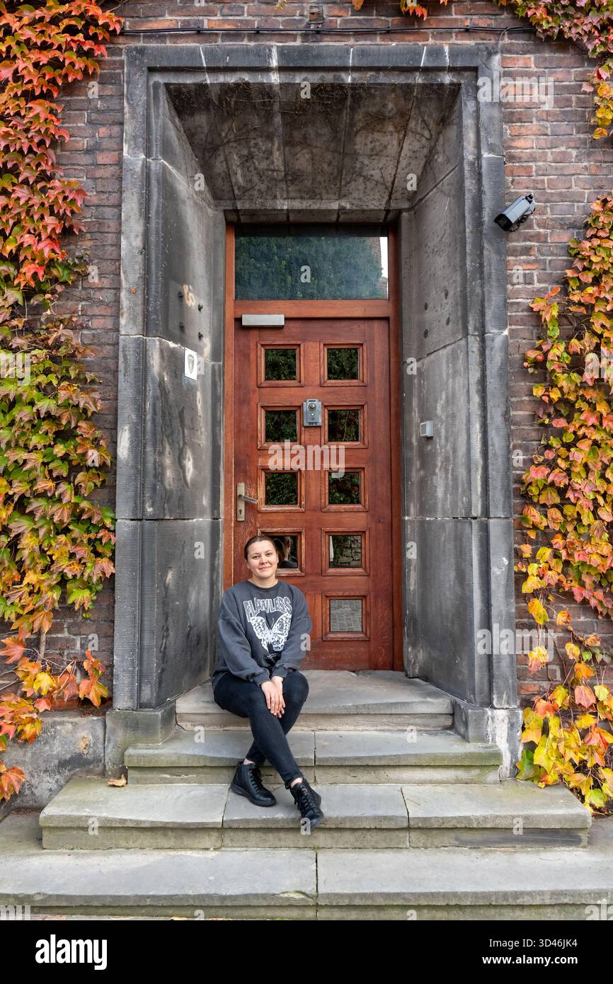 Jeune femme assise sur des marches en pierre devant l'entrée du bâtiment historique orné avec lierre d'automne coloré couvrant des murs de briques. Concept d'autum urbain Banque D'Images