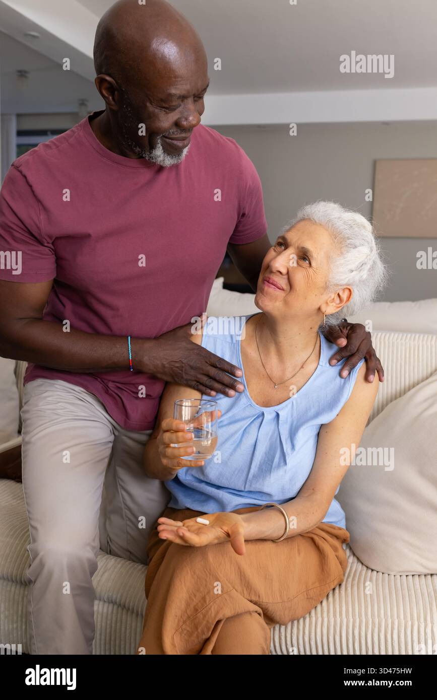 Couple senior diversifié partageant moment dans le salon, avec canapé, pilule et verre d'eau Banque D'Images