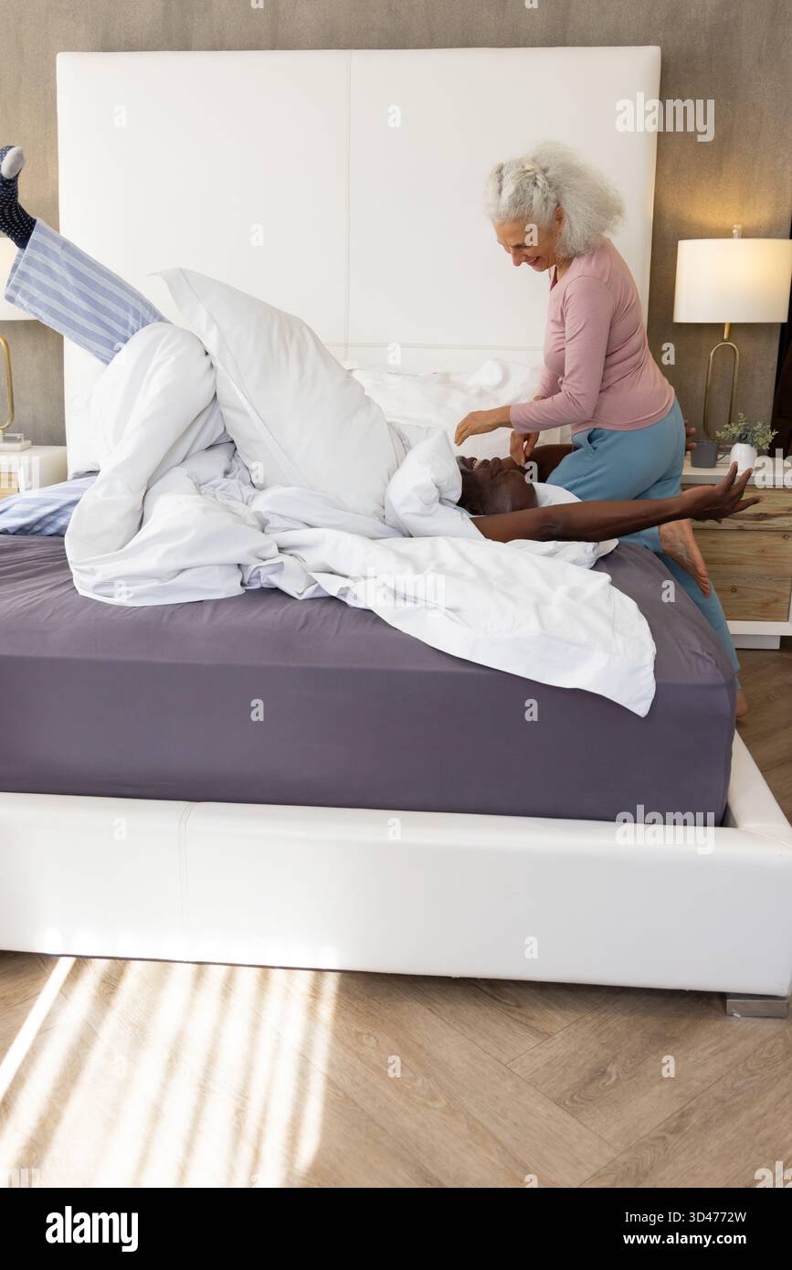 Couple senior diversifié s'étirant ensemble sur le lit dans une chambre moderne, avec lampe de chevet et plante Banque D'Images