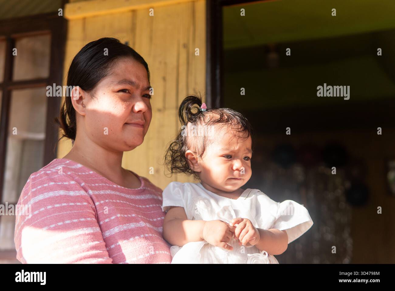 Un portrait franc d'une jolie petite fille asiatique dans une robe blanche, assise avec sa mère. Les deux regardent loin réfléchi sur un porche ensoleillé dans une maison chaude. Banque D'Images