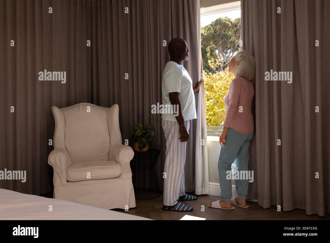 Debout couple senior diversifié tirant les rideaux gris à la fenêtre de la chambre, avec des plantes en pot Banque D'Images