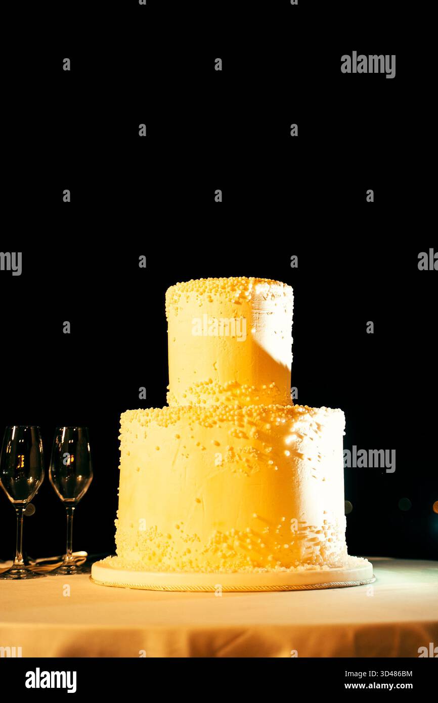 Gâteau de mariage à deux niveaux élégamment décoré de perles, reposant sur une table avec trois verres à vin à côté, sur un fond sombre, créant un Banque D'Images