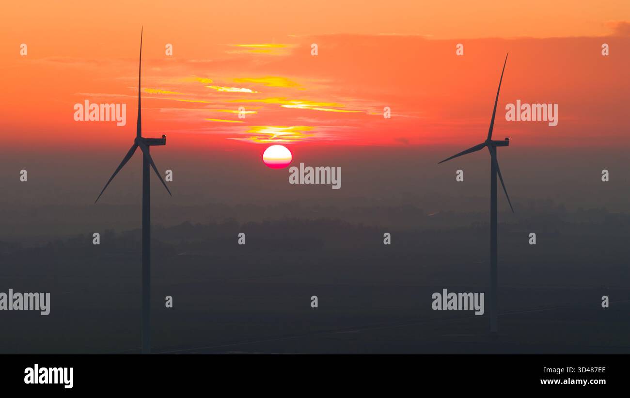 Éoliennes au coucher du soleil au-dessus d'un paysage rural brumeux. Le ciel orange et rouge profond crée une scène dramatique et atmosphérique mettant en valeur les énergies renouvelables Banque D'Images