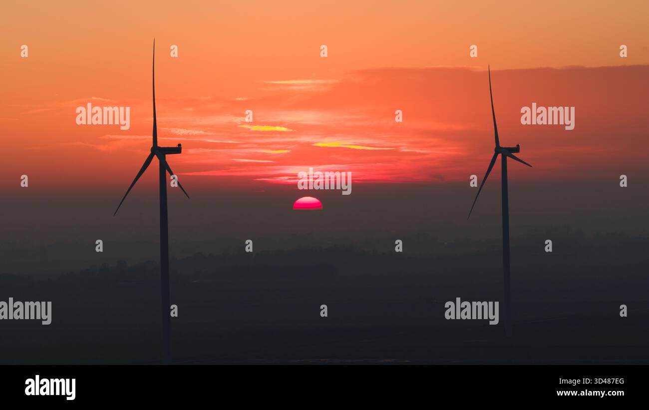 Éoliennes au coucher du soleil au-dessus d'un paysage rural brumeux. Le ciel orange et rouge profond crée une scène dramatique et atmosphérique mettant en valeur les énergies renouvelables Banque D'Images