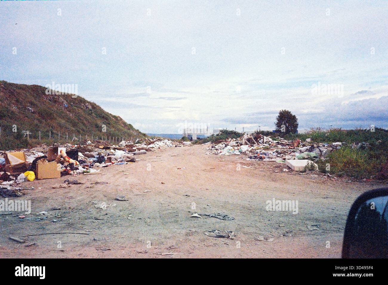 Route passant par une décharge avec des déchets empilés le long, supermarché lointain en vue, environnement urbain brut capturé de style analogique. Banque D'Images