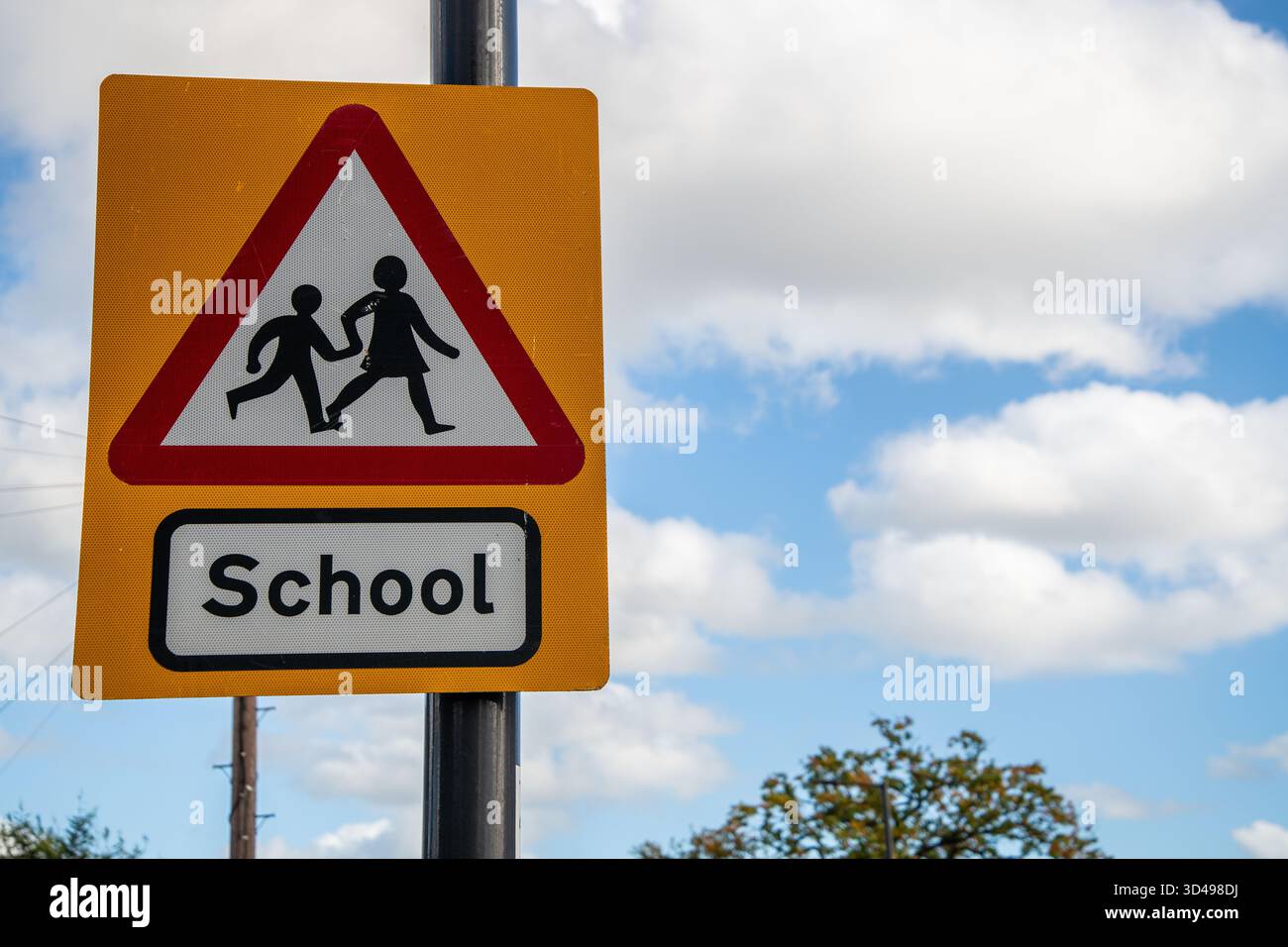 Triangle panneau d'avertissement scolaire sur fond jaune à côté d'une route britannique, alertant les conducteurs d'une école voisine et des enfants traversant. Banque D'Images