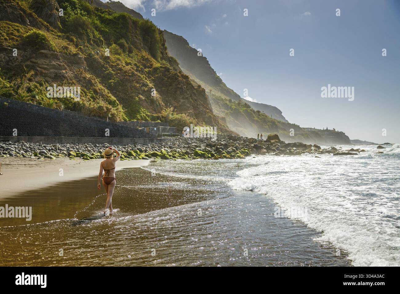 Femme dans un chapeau de paille marche le long d'une Playa del Socorro à Tenerife à côté de falaises accidentées, profitant du temps ensoleillé. Les vagues tournent doucement sur le rivage comme sh Banque D'Images