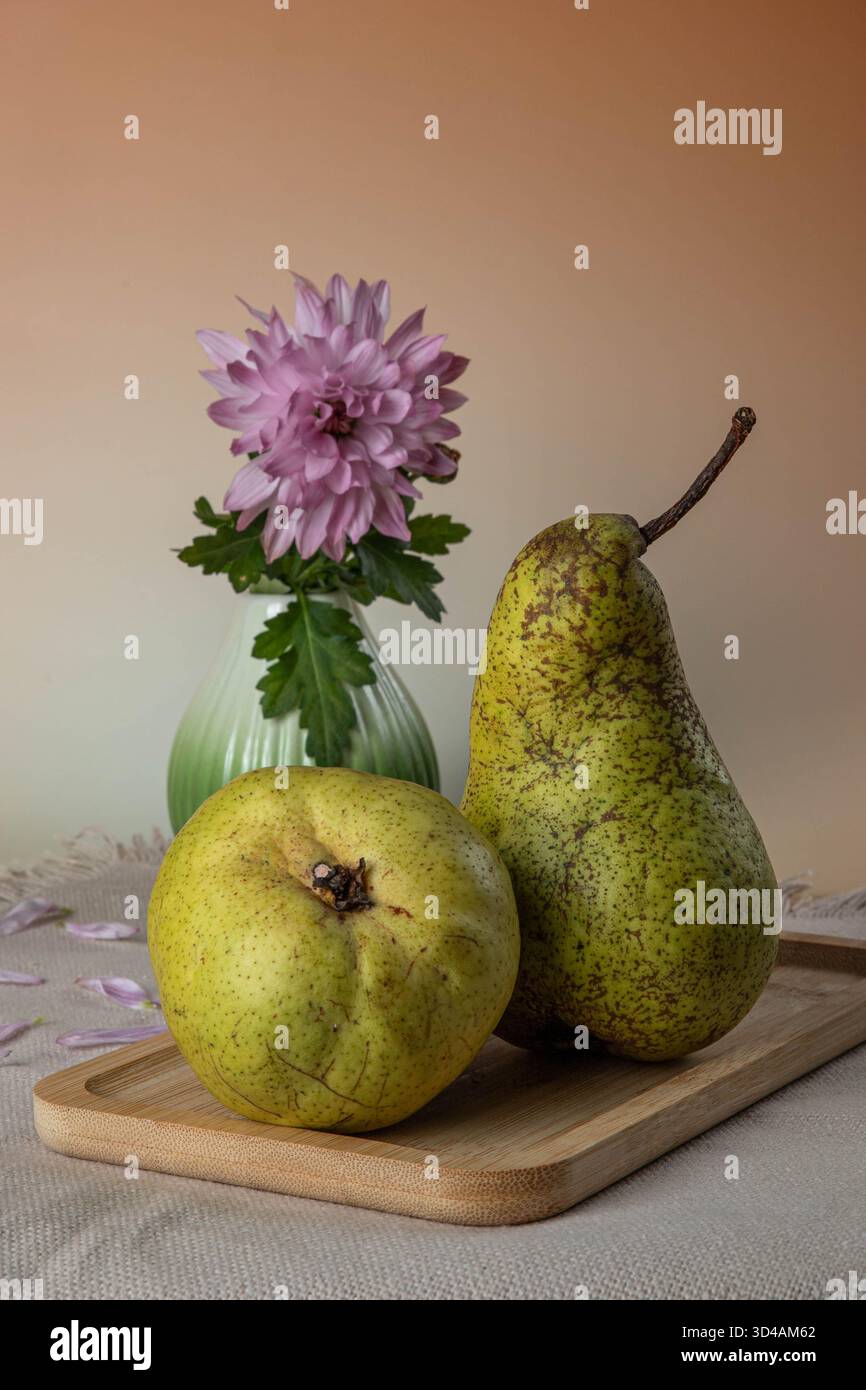 Une poire et un coing reposent sur une planche à découper en bois. À proximité, un vase côtelé vert contient un chrysanthème rose (Chrysanthemum morifolium) et une feuille verte Banque D'Images