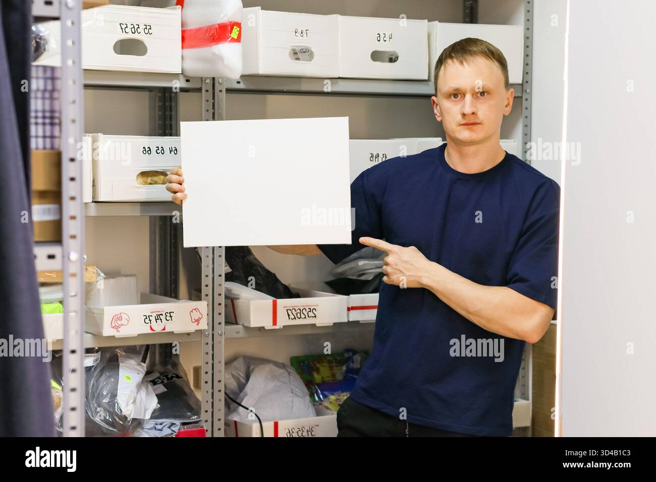 Homme tenant une feuille vierge et pointant dans le stockage. L'employé pointe en tenant du papier blanc devant les étagères contenant des marchandises entreposées Banque D'Images