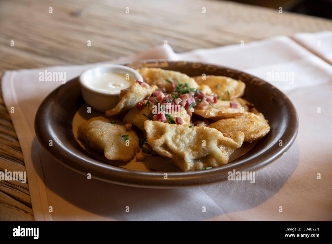 Piérogies polonaises classiques avec garniture de jambon et d'oignons verts, servies sur une assiette en faïence brune posée sur une serviette en lin, accompagnée d'une table en bois. Banque D'Images