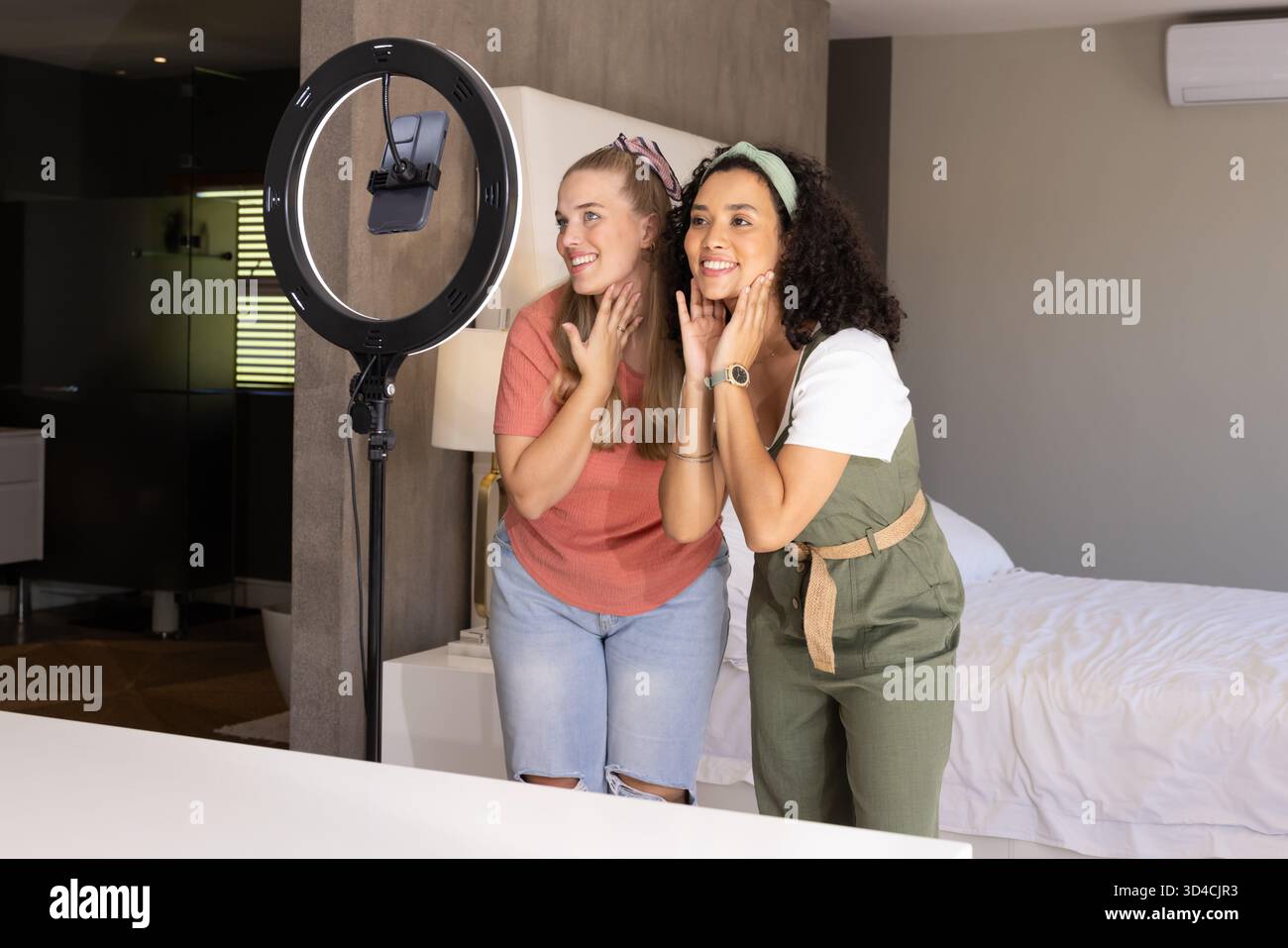 Des amies féminines souriantes diverses démontrant des soins de la peau dans la chambre, avec lumière annulaire et smartphone Banque D'Images