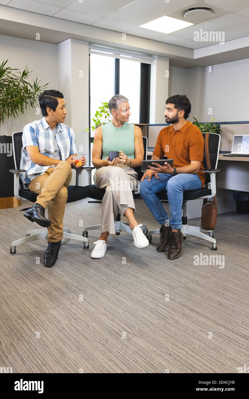 Divers collègues assis dans un bureau ouvert discutant du projet, tenant des tasses à café et une tablette Banque D'Images