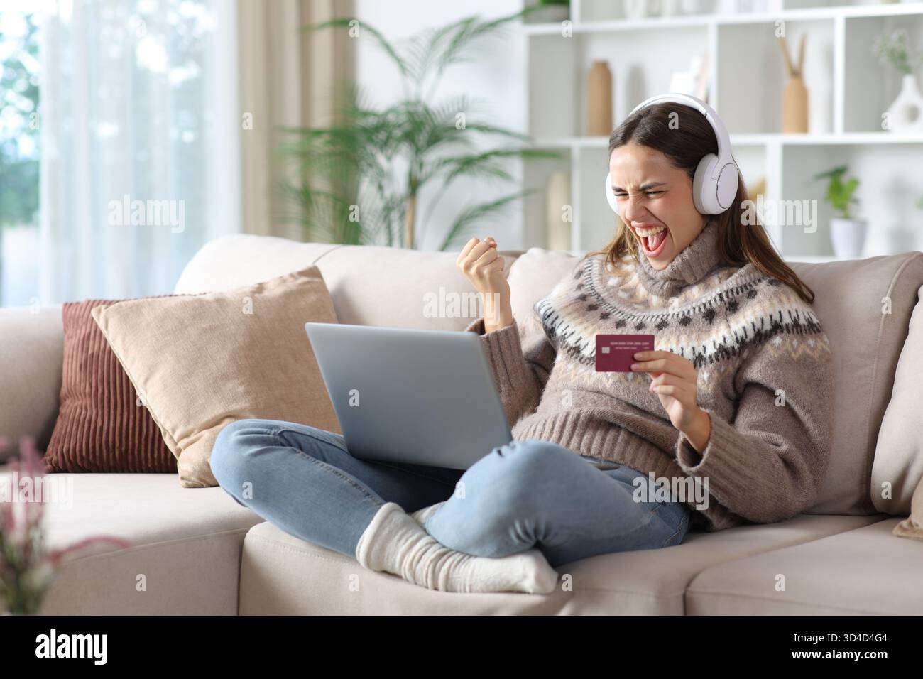 Femme excitée en hiver achetant des médias en ligne en utilisant la carte de crédit et ordinateur portable assis sur un canapé à la maison Banque D'Images