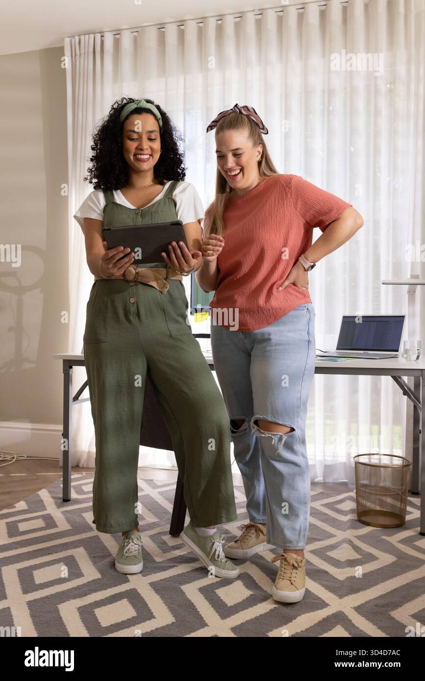 Tenant la tablette, diverses collègues femmes debout côte à côte dans le bureau à domicile, examinant le contenu Banque D'Images
