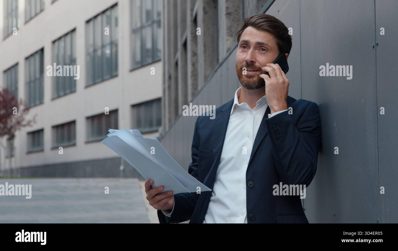 Homme d'affaires d'âge moyen caucasien homme d'affaires parlant téléphone tenant des papiers dans la ville penchant mur de bureau homme d'affaires masculin mature debout dedans Banque D'Images