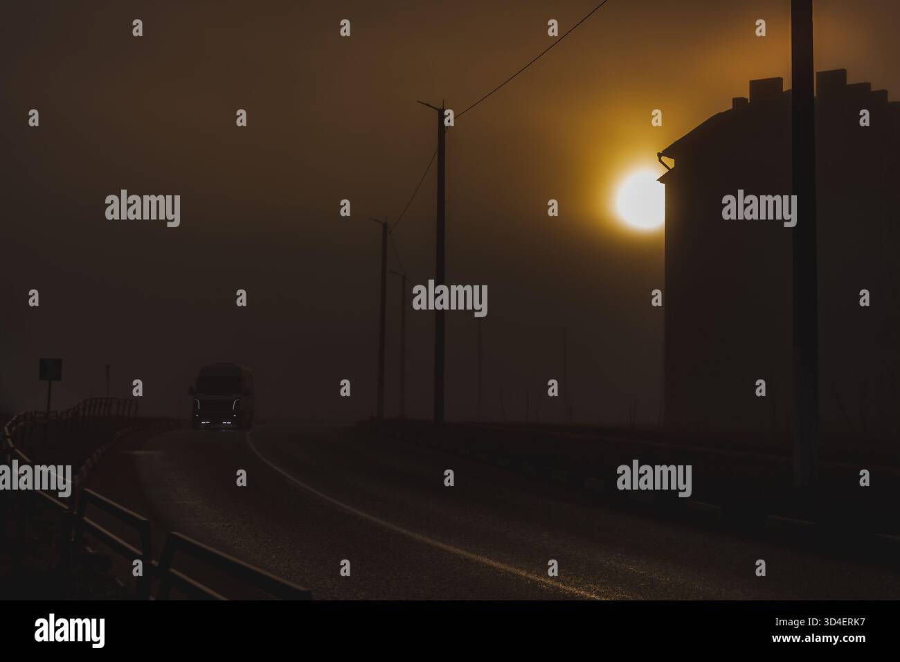 Un camion solitaire traverse une route brumeuse au lever du soleil. La lumière dorée perce le brouillard, créant une ambiance cinématographique et paisible. Banque D'Images