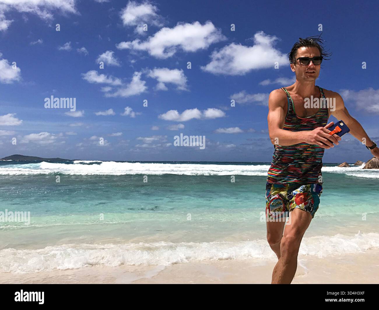 Bel homme sportif en tenue d'été courant sur la plage profitant du paysage turquoise de l'océan Indien, tout en tenant le téléphone à la main. Modèle masculin de mode Banque D'Images