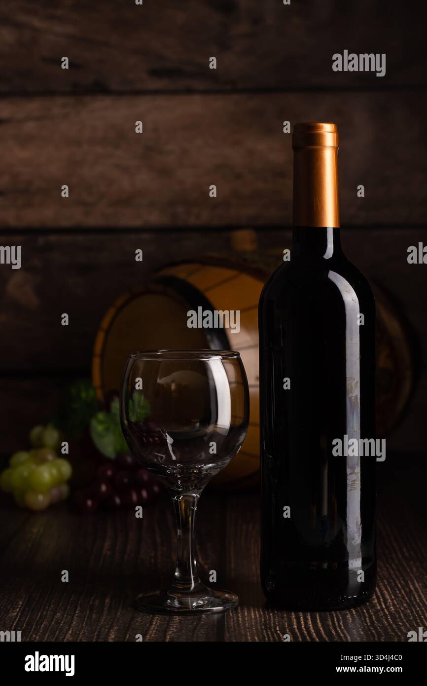 Une bouteille de vin se tient haute à côté d'un verre vide sur une table en bois. Les raisins entourent les éléments, avec un tonneau en arrière-plan, créant une atmosphère confortable Banque D'Images