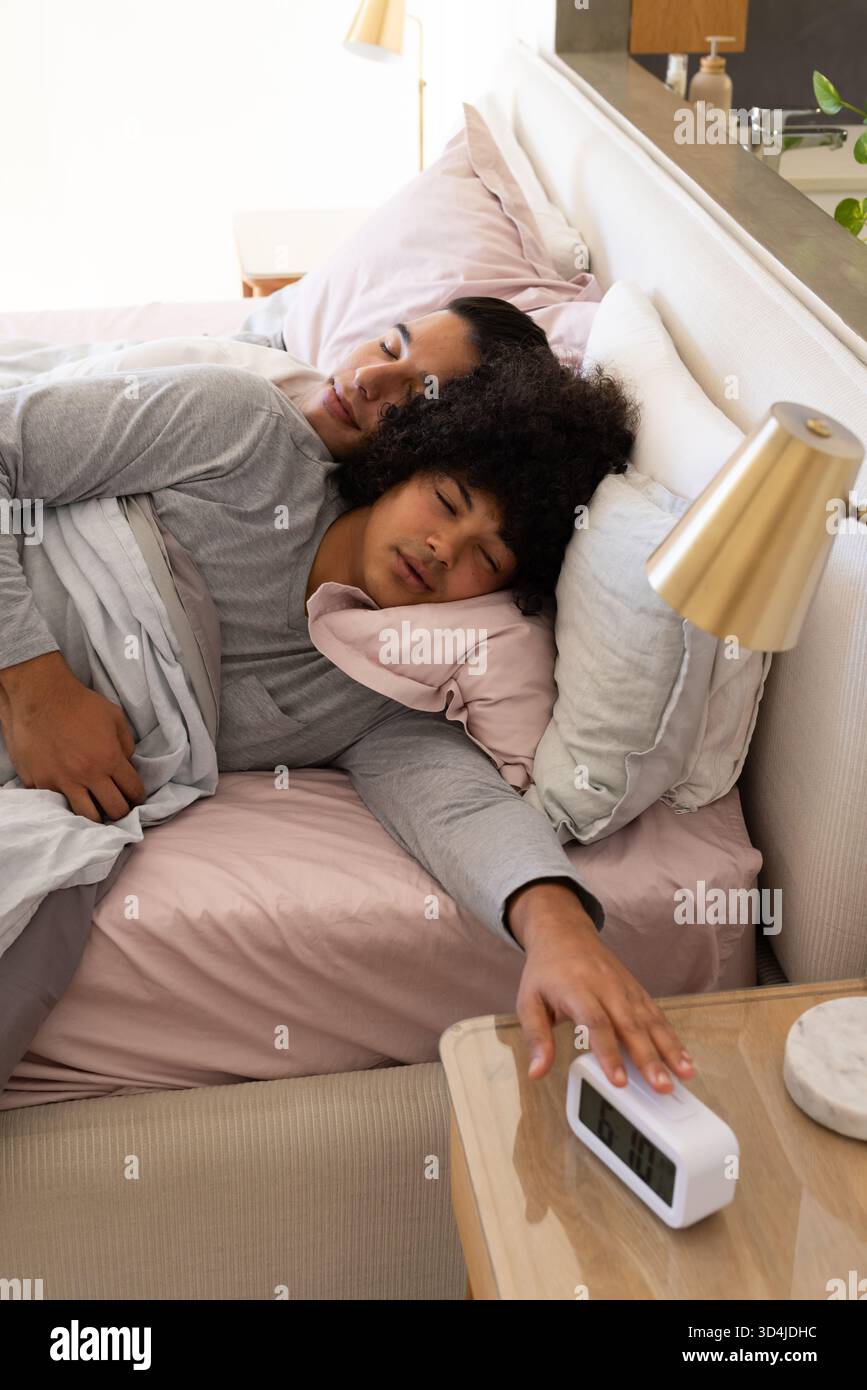 Dormir divers partenaires masculins atteignant vers le réveil sur la table de nuit dans la chambre, avec des oreillers Banque D'Images