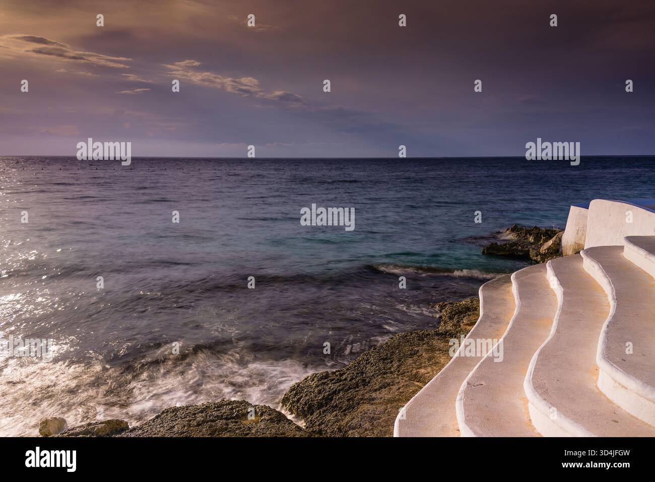 Une photographie horizontale à longue exposition d'escaliers en béton blancs et ondulés sur une côte rocheuse, avec des vagues floues et un ciel crépusculaire sombre et violet. Banque D'Images