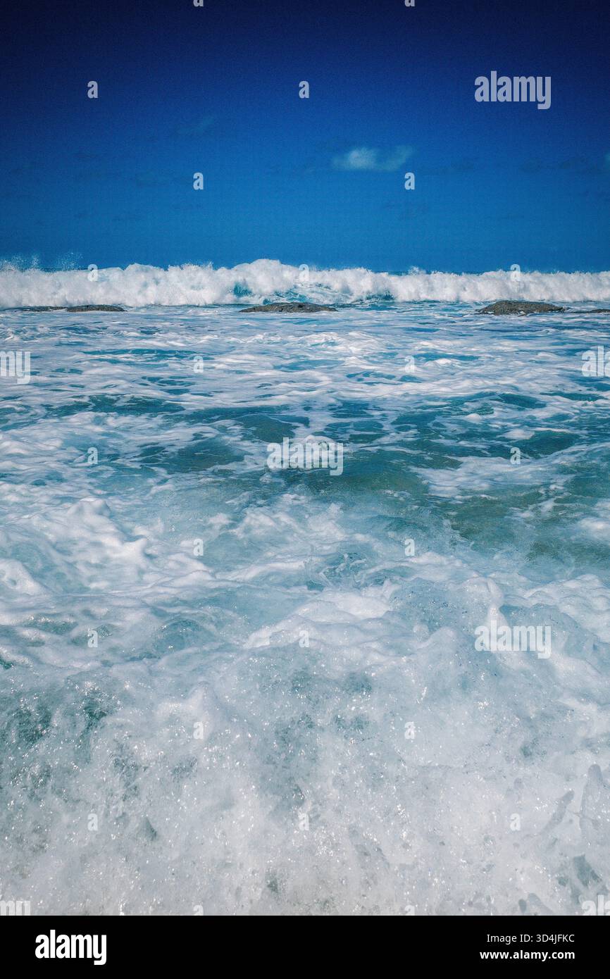 Une vue verticale, à hauteur des yeux, des vagues blanches mousseuses de l'océan et du surf sous un ciel bleu profond et granuleux. Banque D'Images