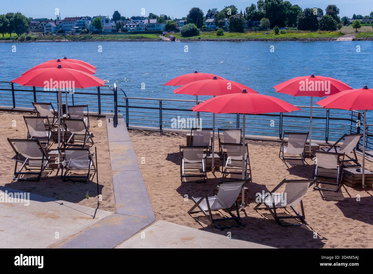 Une plage au bord du Rhin est agrémentée de rangées de parasols rouges et de chaises longues sur du sable mou. La scène capture une journée d'été sereine avec clair b Banque D'Images