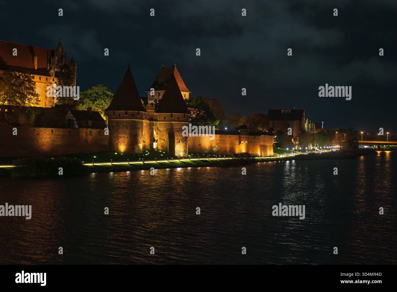 Les murs de briques rouges et les tours coniques brillent de lumières chaudes après le crépuscule. Les reflets ondulent sur la rivière Nogat alors que la vaste forteresse gothique domine le s. Banque D'Images