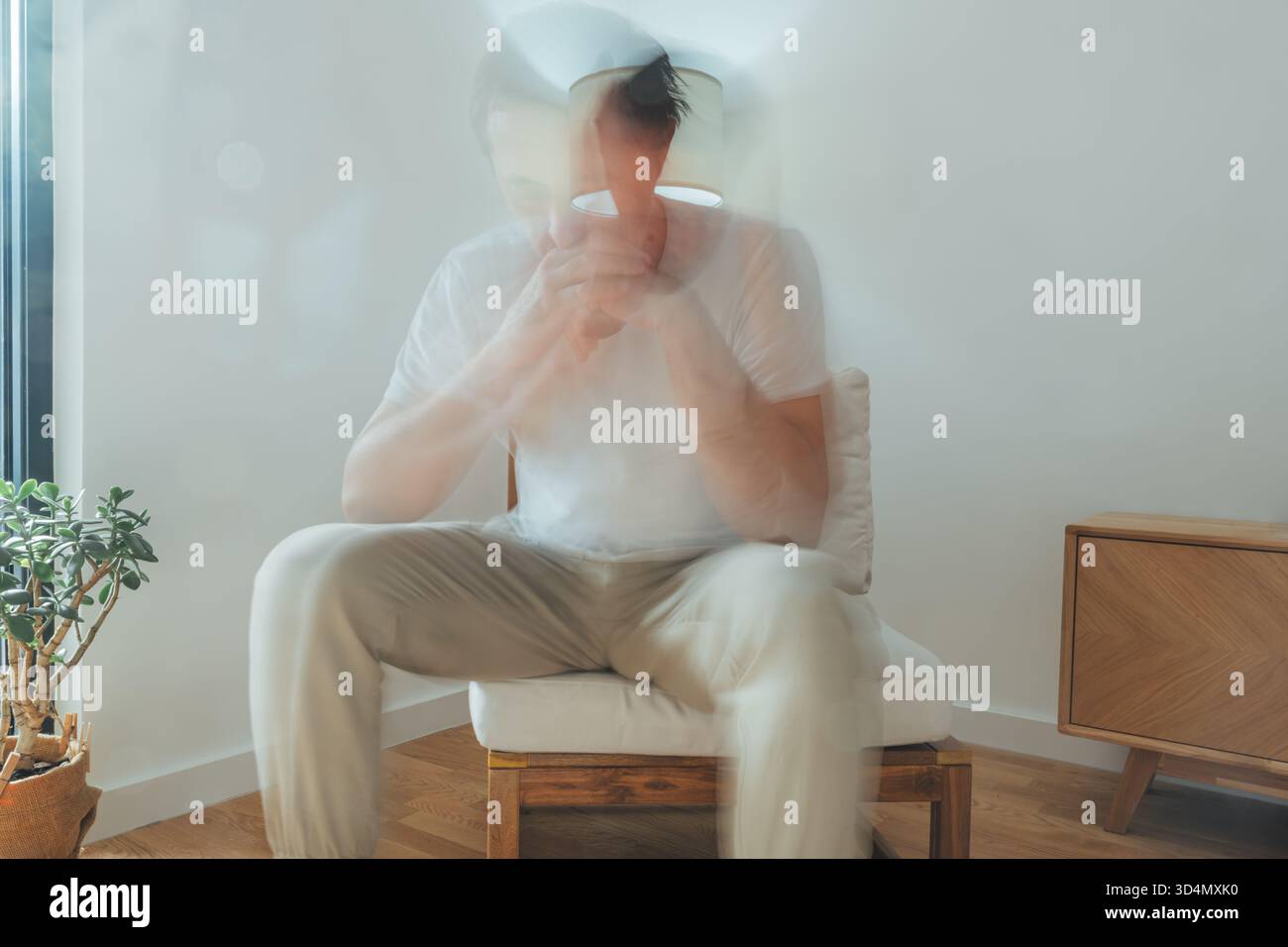 Homme mûr assis dans une chaise avec un mouvement flou, les mains serrées devant le visage, montrant le stress et la lutte émotionnelle dans un intérieur calme de la maison. Sel Banque D'Images