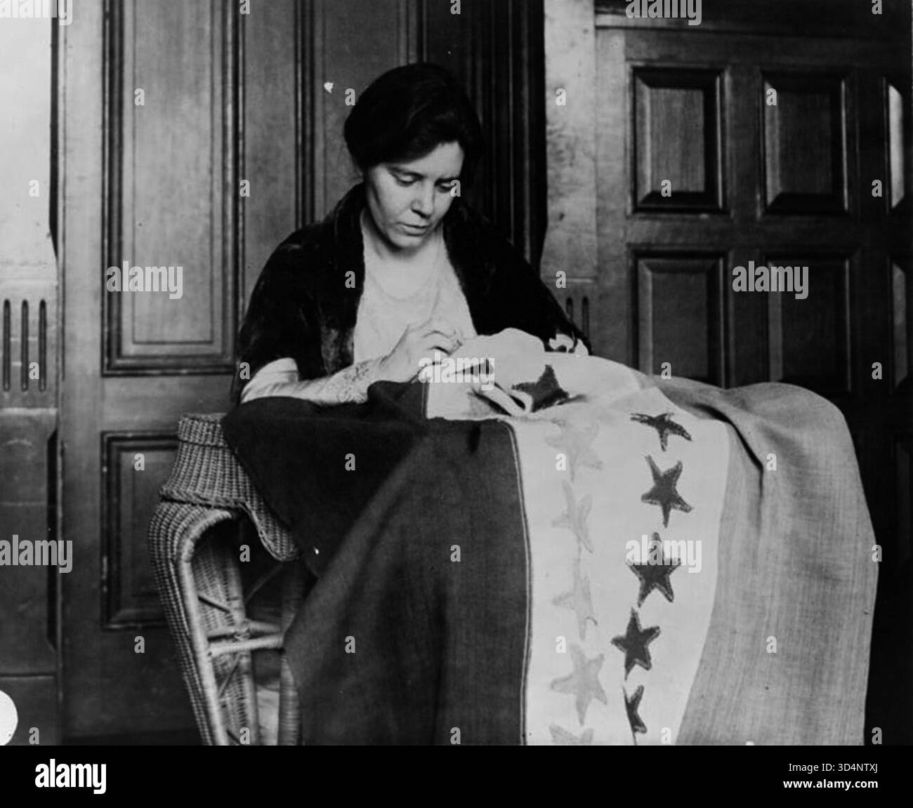 Portrait en demi-longueur d'Alice Paul, assise et légèrement tournée vers la droite, cousant un drapeau de suffrage. La photographie capture Paul, une suffragette américaine de premier plan et stratège du National Woman’s Party, engagée dans un travail d’activisme pour le droit de vote des femmes au début du XXe siècle. Banque D'Images
