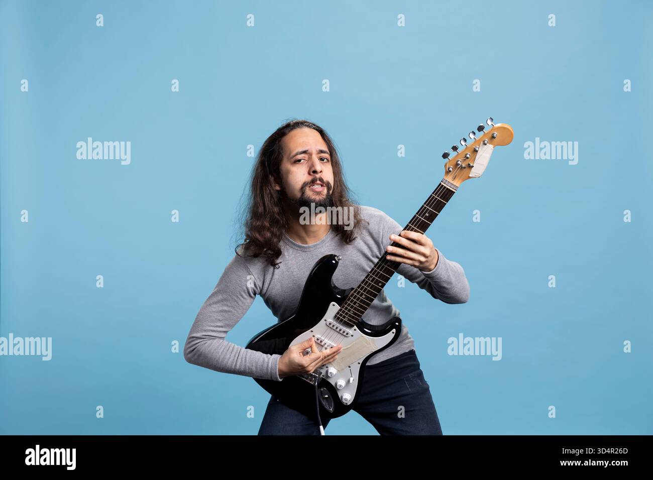 Jeune guitariste masculin aux cheveux longs se produisant passionnément sur une guitare électrique noire dans une pose dynamique. Musicien énergique respire les vibrations rock star, la joie et l'enthousiasme sur un fond de studio. Banque D'Images