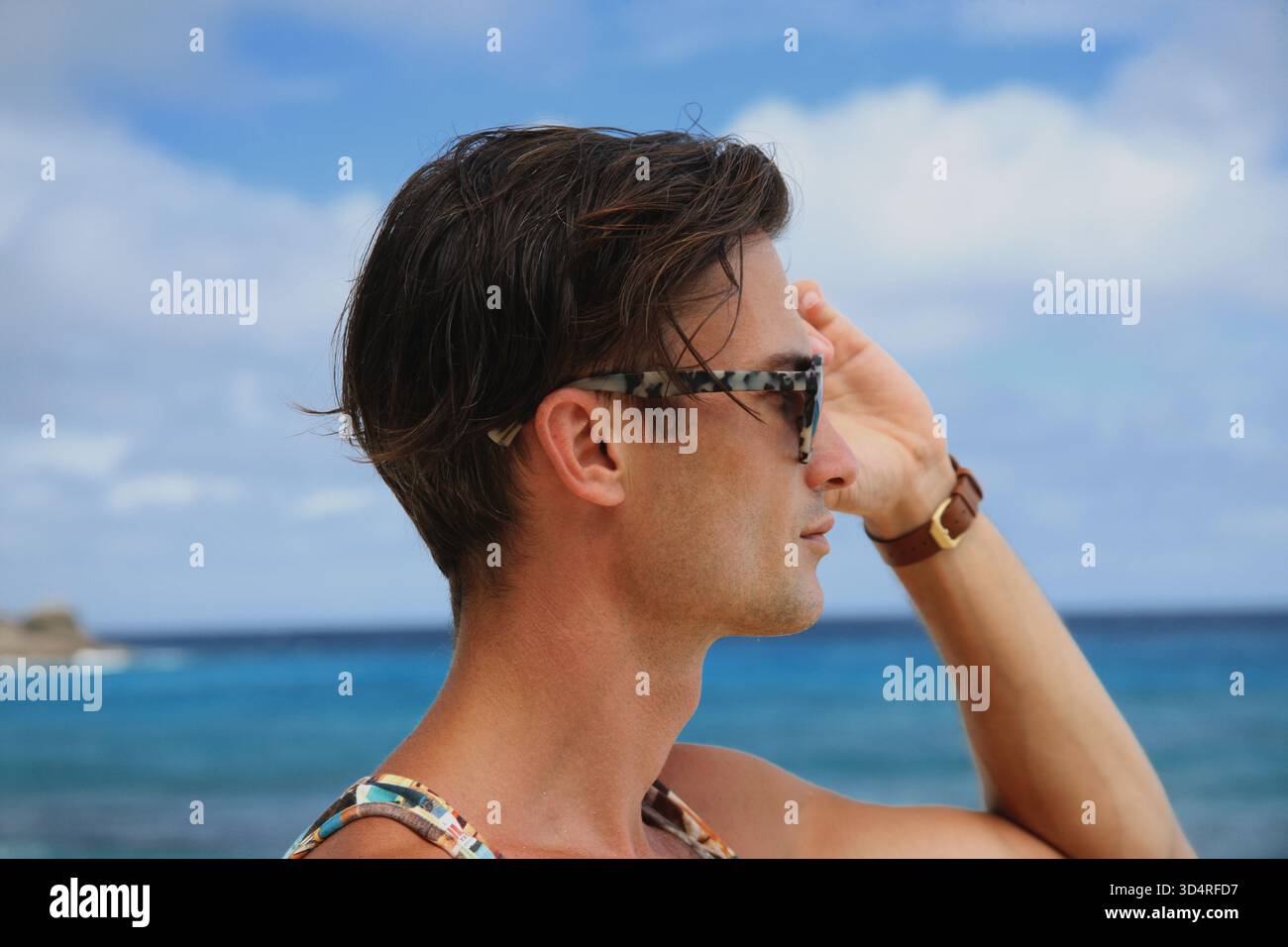 Photo de tête d'un beau mannequin masculin regardant latéralement à travers des lunettes de soleil sur une plage tropicale. Portrait de modèle masculin avec vue mer turquoise sur fond. Banque D'Images