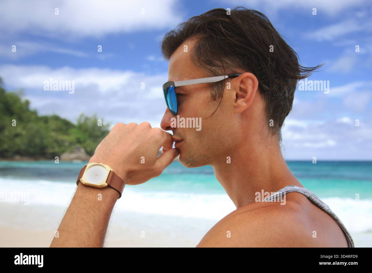Photo de tête d'un beau mannequin masculin regardant latéralement à travers des lunettes de soleil sur une plage tropicale. Portrait de modèle masculin avec vue mer turquoise sur fond. Banque D'Images