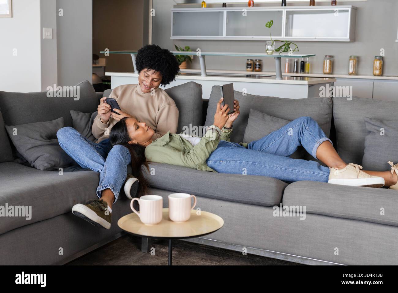 Se prélasser couple diversifié sur le canapé dans le salon ouvert, la navigation smartphone et tablette Banque D'Images