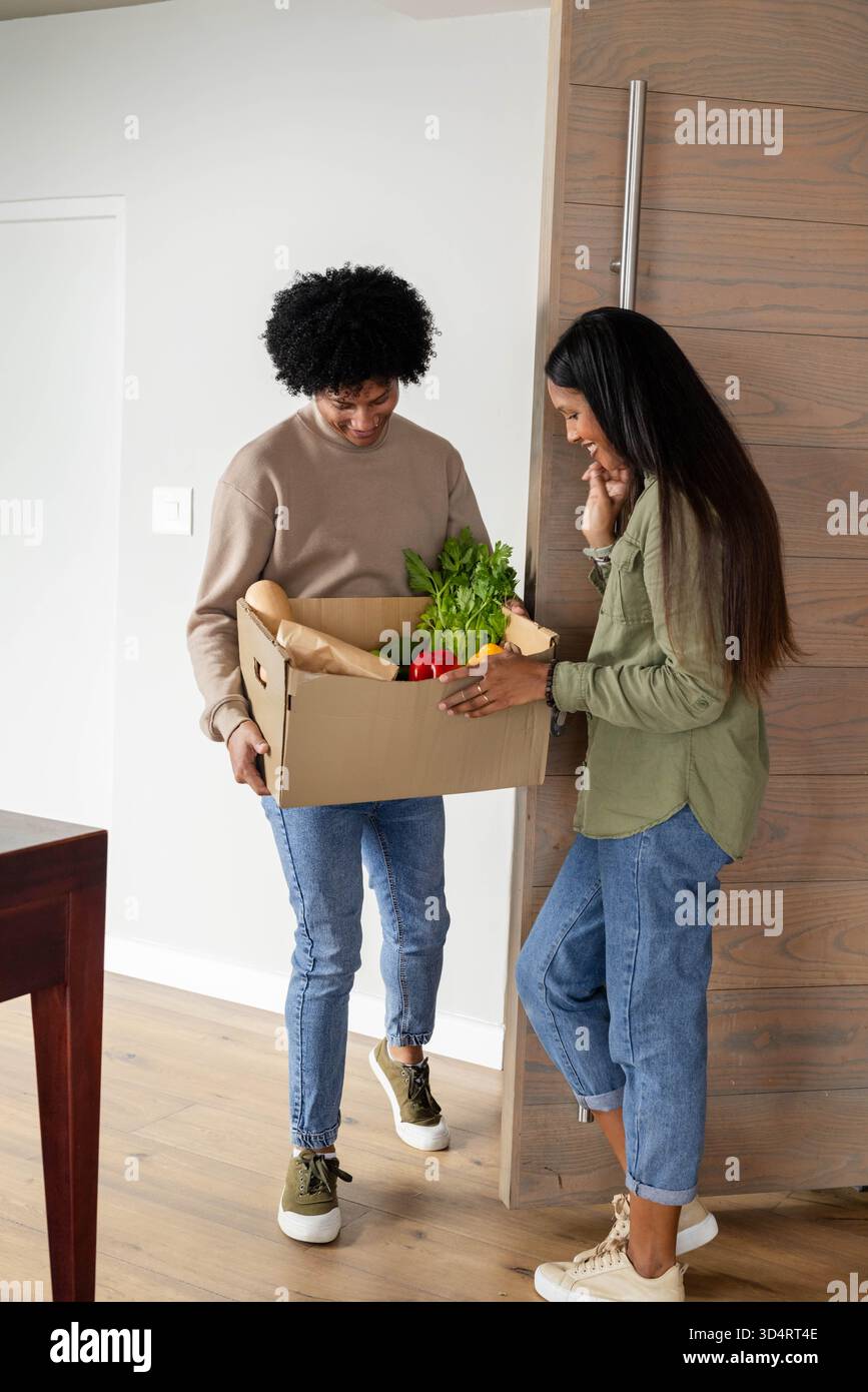 Debout couple diversifié tenant l'épicerie à l'entrée de la maison, avec boîte en carton de produits frais Banque D'Images