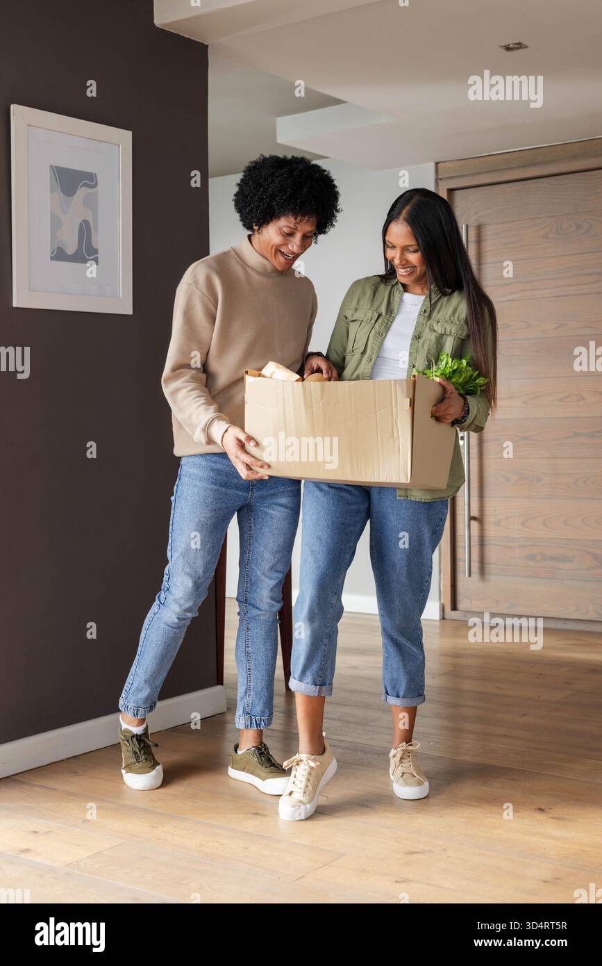 Inspecter la boîte d'épicerie en carton, couple diversifié debout dans l'entrée de la maison avec du pain et des légumes verts Banque D'Images