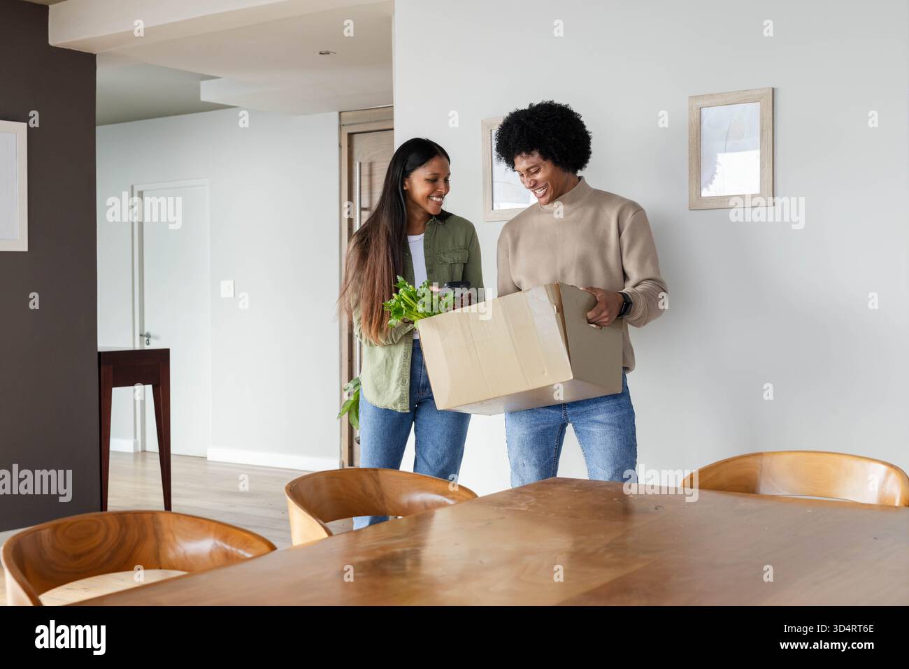 Déballage d'un couple diversifié tenant une boîte en carton avec des verts feuillus frais dans la salle à manger moderne Banque D'Images