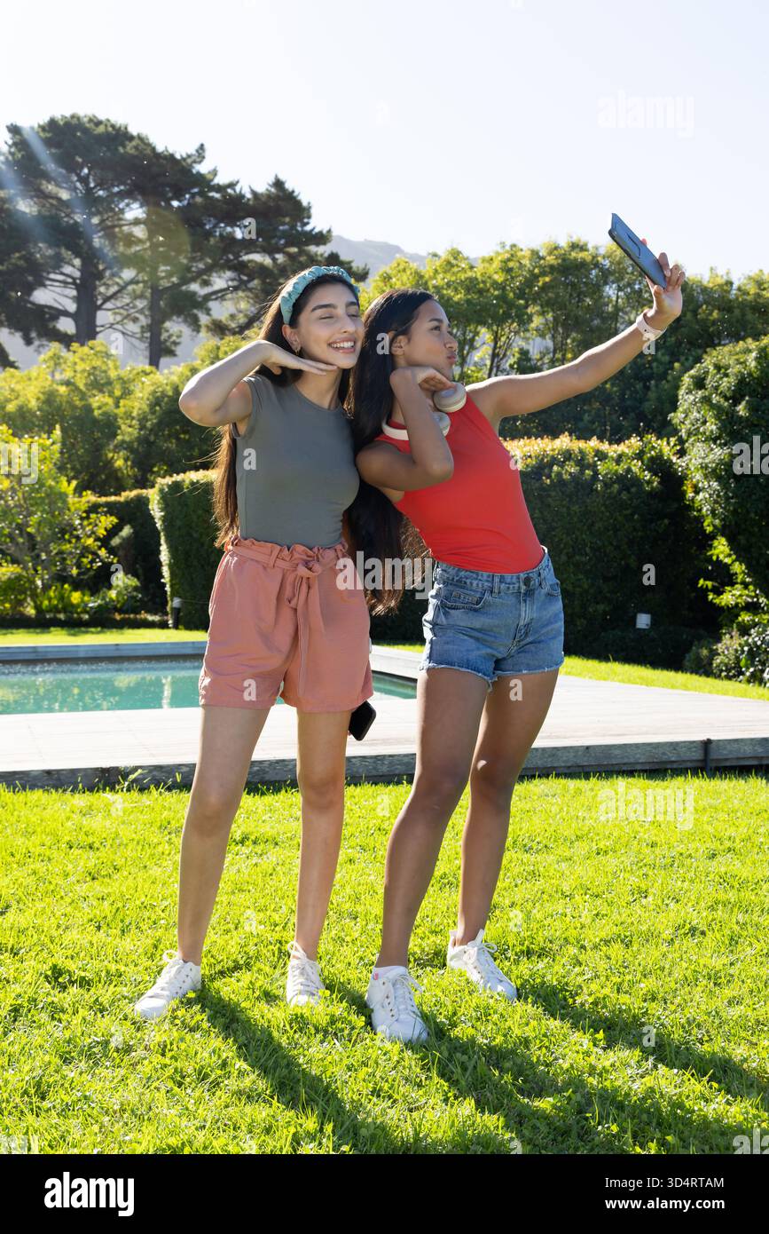 Prise selfie diverses amies féminines posant sur la pelouse luxuriante de la cour à côté de la piscine, avec smartphone Banque D'Images