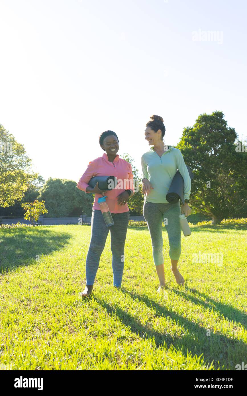 Marchant diverses amies discutant sur la pelouse du parc, portant des tapis de yoga et des bouteilles d'eau Banque D'Images