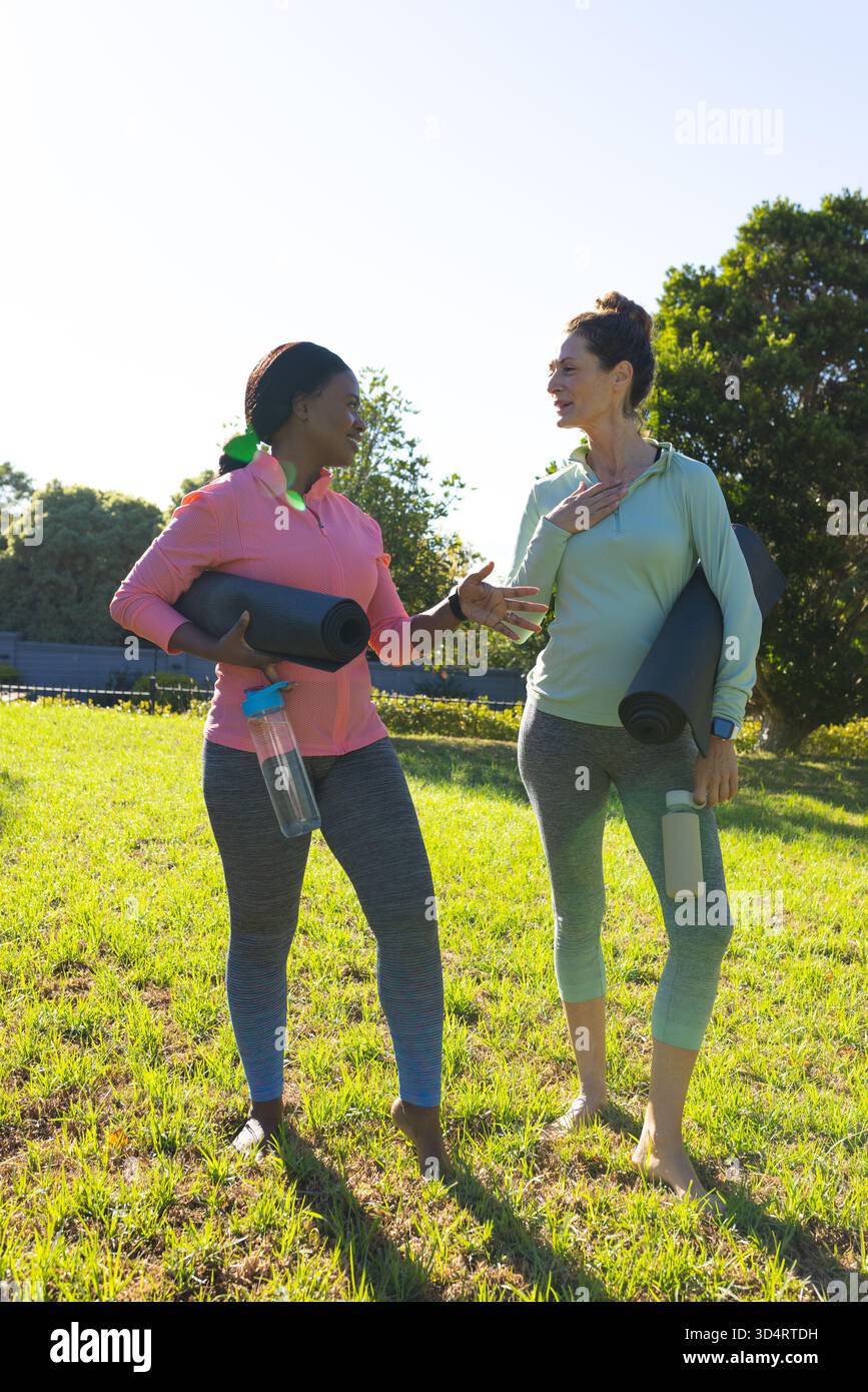 Bavardez diverses amies féminines tenant des tapis de yoga et des bouteilles d'eau sur un terrain herbeux avec des tapis noirs Banque D'Images