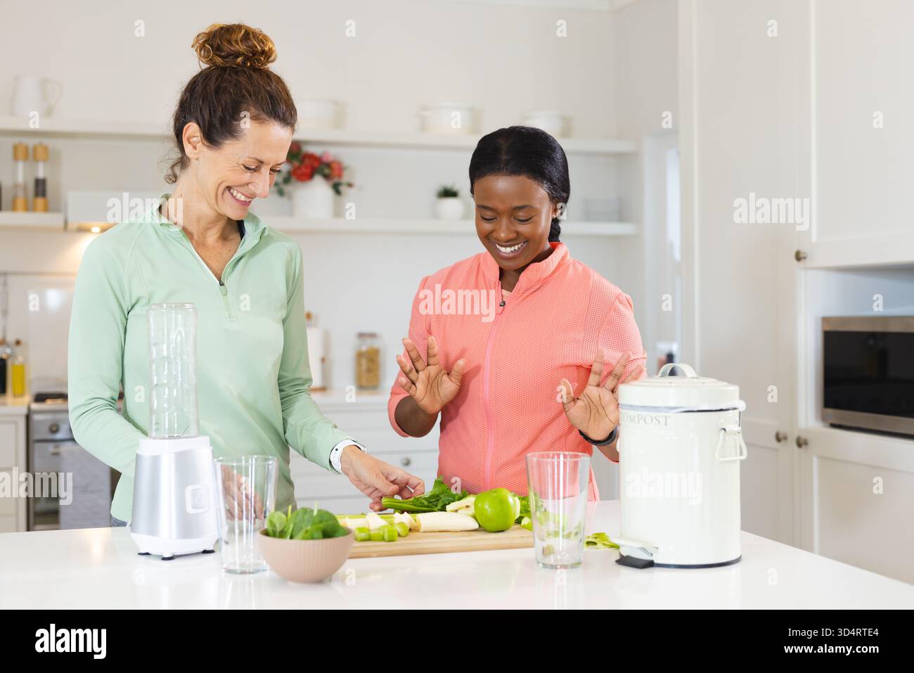 Amies féminines diverses coupant le chou frisé et les pommes dans la cuisine à la maison, avec base de mélangeur et bac à compost Banque D'Images
