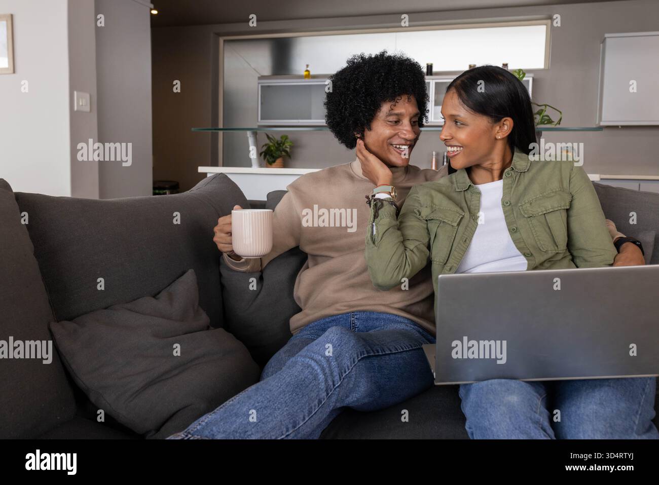 Couple diversifié tenant une tasse en céramique et utilisant un ordinateur portable sur un canapé dans le salon, avec des oreillers de jet Banque D'Images