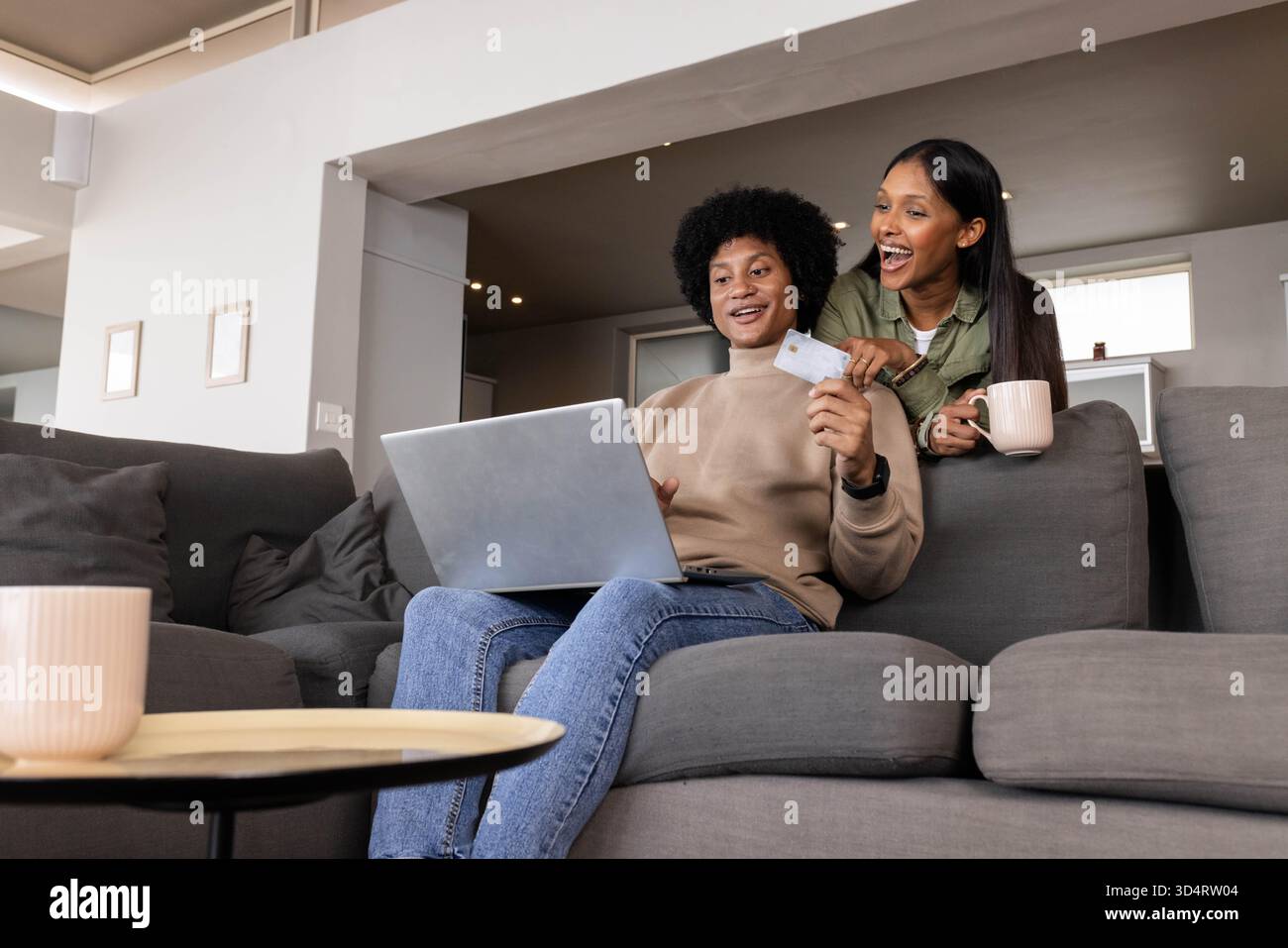 Shopping en ligne couple diversifié assis dans le salon moderne, avec tasse à café de carte de crédit d'ordinateur portable Banque D'Images