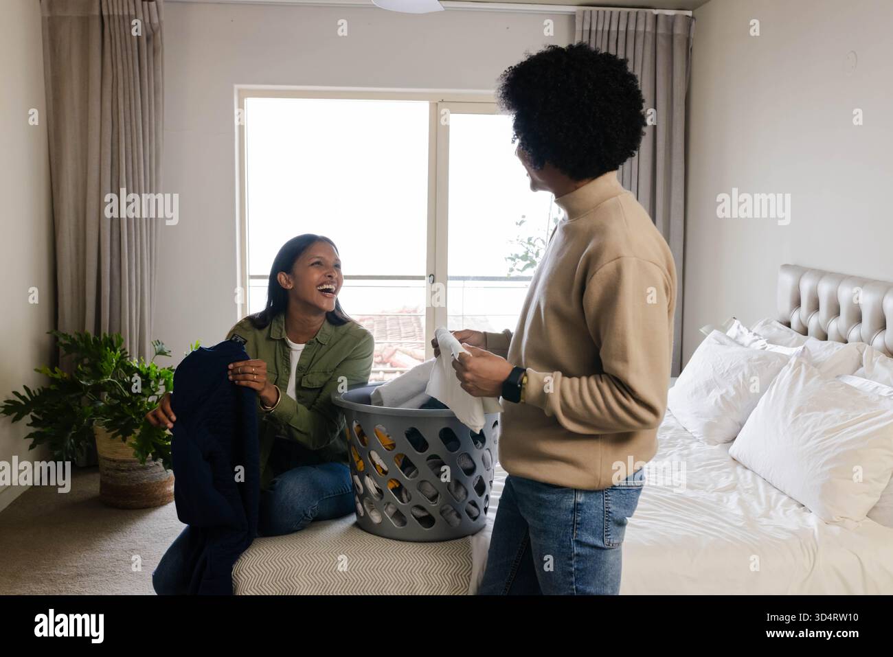 Couple diversifié pliant le linge ensemble dans la chambre à coucher à côté de la plante en pot, avec panier à linge gris Banque D'Images