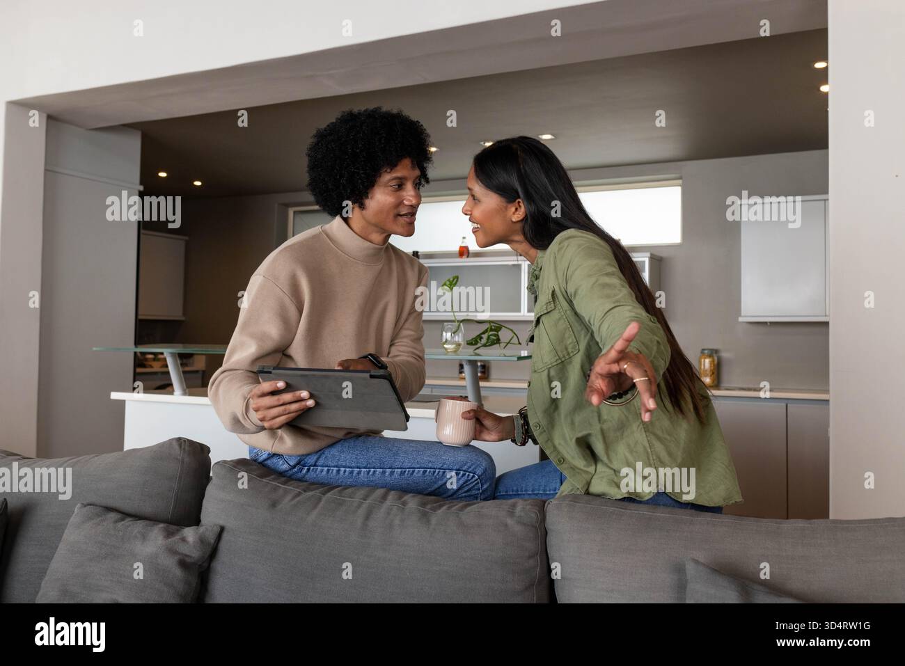 Discuter couple diversifié assis sur le canapé dos dans un espace de vie ouvert, avec tablette et tasse Banque D'Images