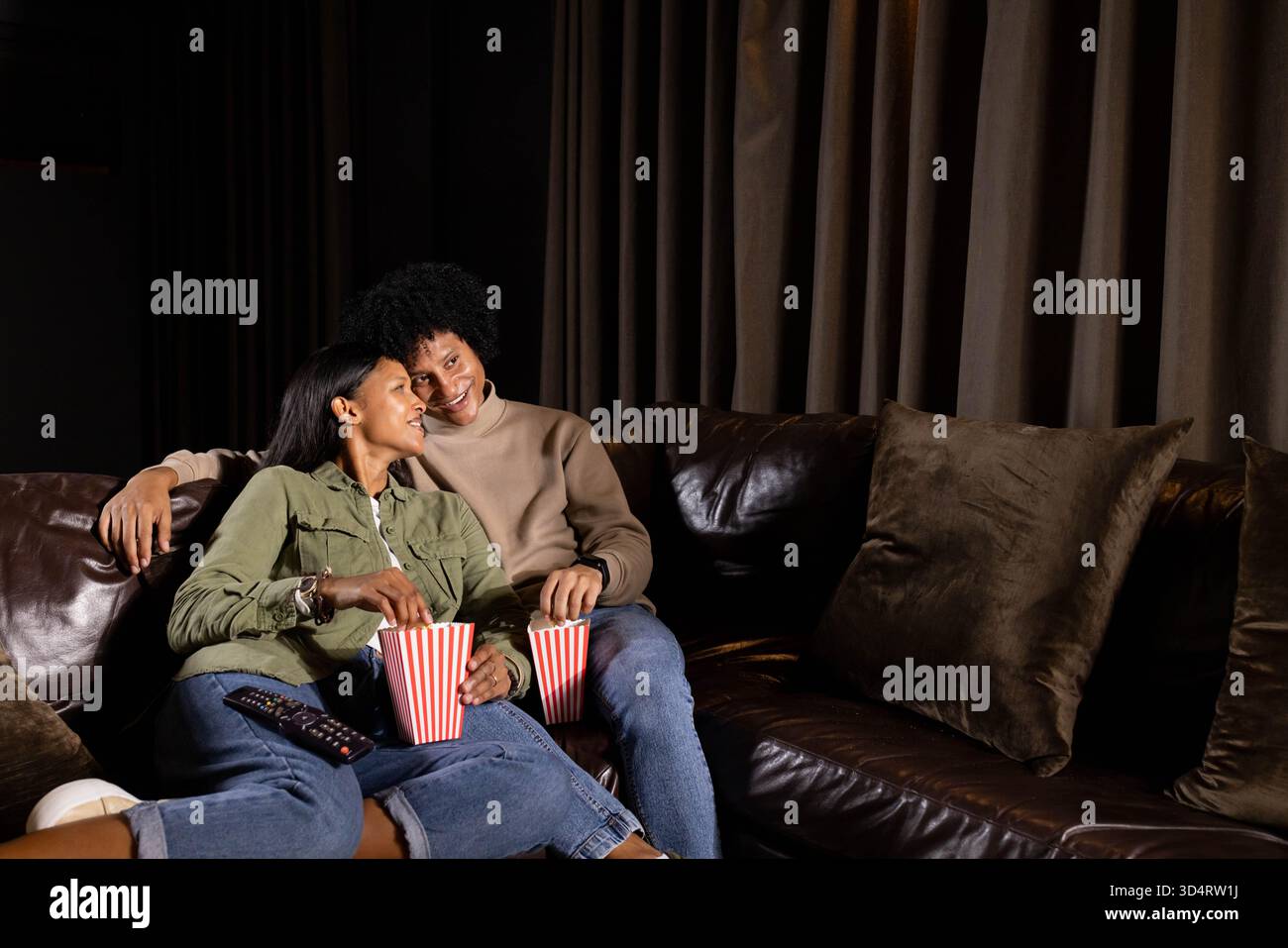 Couple adulte moyen diversifié appuyé sur un canapé en cuir dans le salon tenant du pop-corn rayé et à distance Banque D'Images