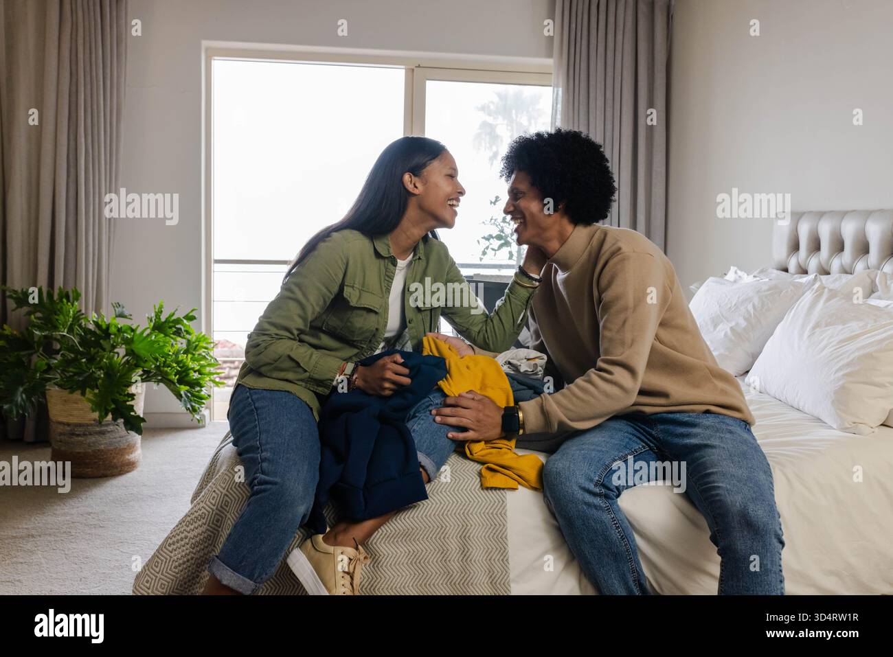 Tri de divers couples manipulant des vêtements pliés sur le lit dans une chambre éclairée, avec des plantes en pot Banque D'Images