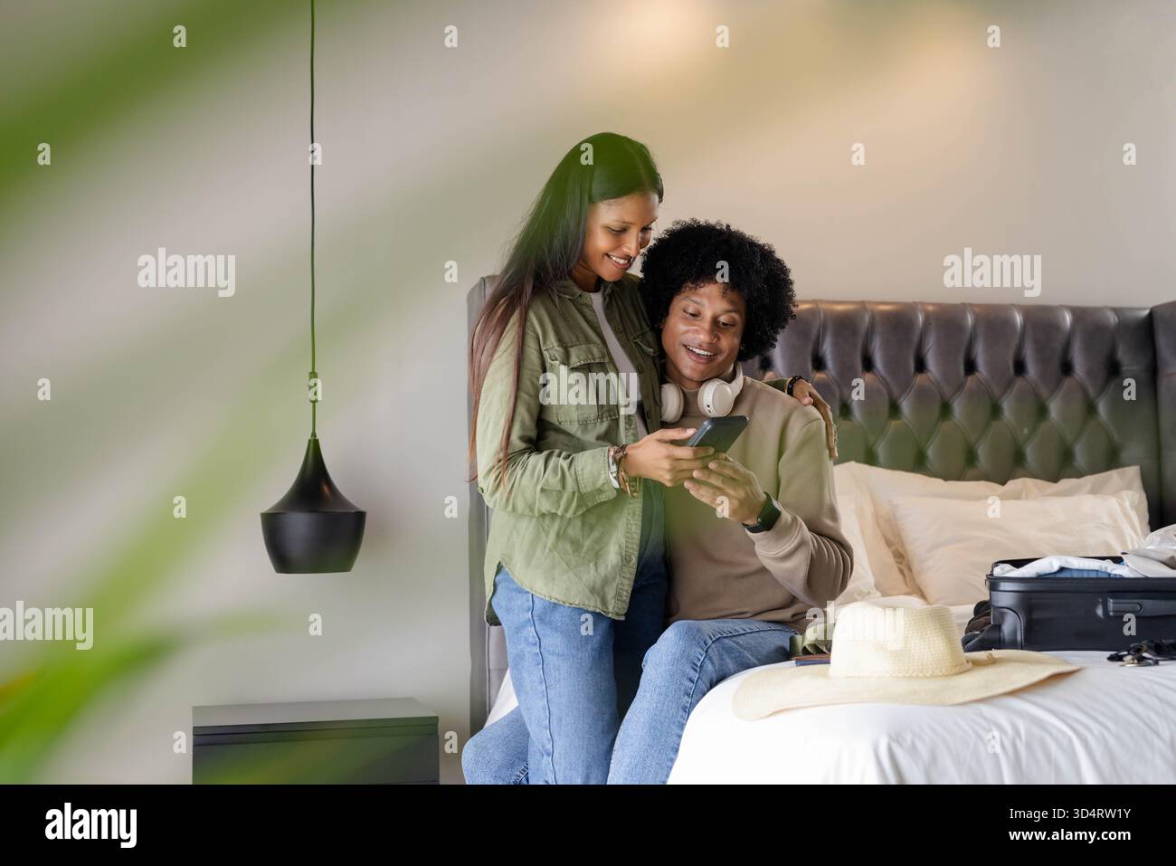 Couple souriant divers assis sur le bord du lit dans la chambre, avec smartphone et valise ouverte Banque D'Images