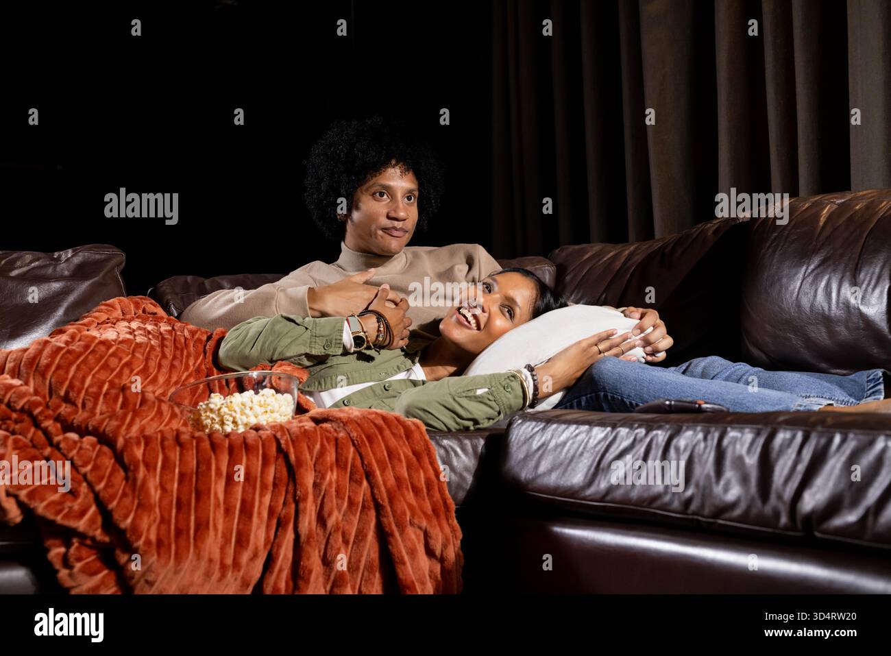 Se prélasser couple diversifié se détendre sur le canapé en cuir dans le salon faiblement éclairé, avec bol à pop-corn Banque D'Images